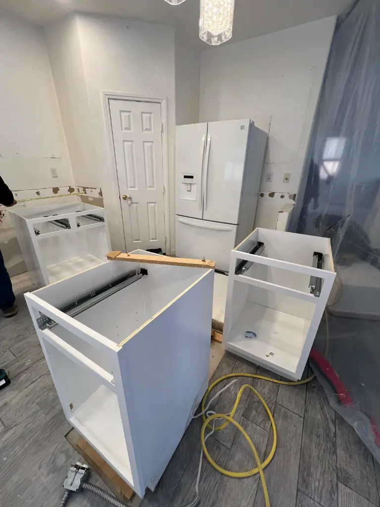 Kitchen renovation in progress with white base cabinets placed on the floor, exposed backsplash area, and a refrigerator against the wall