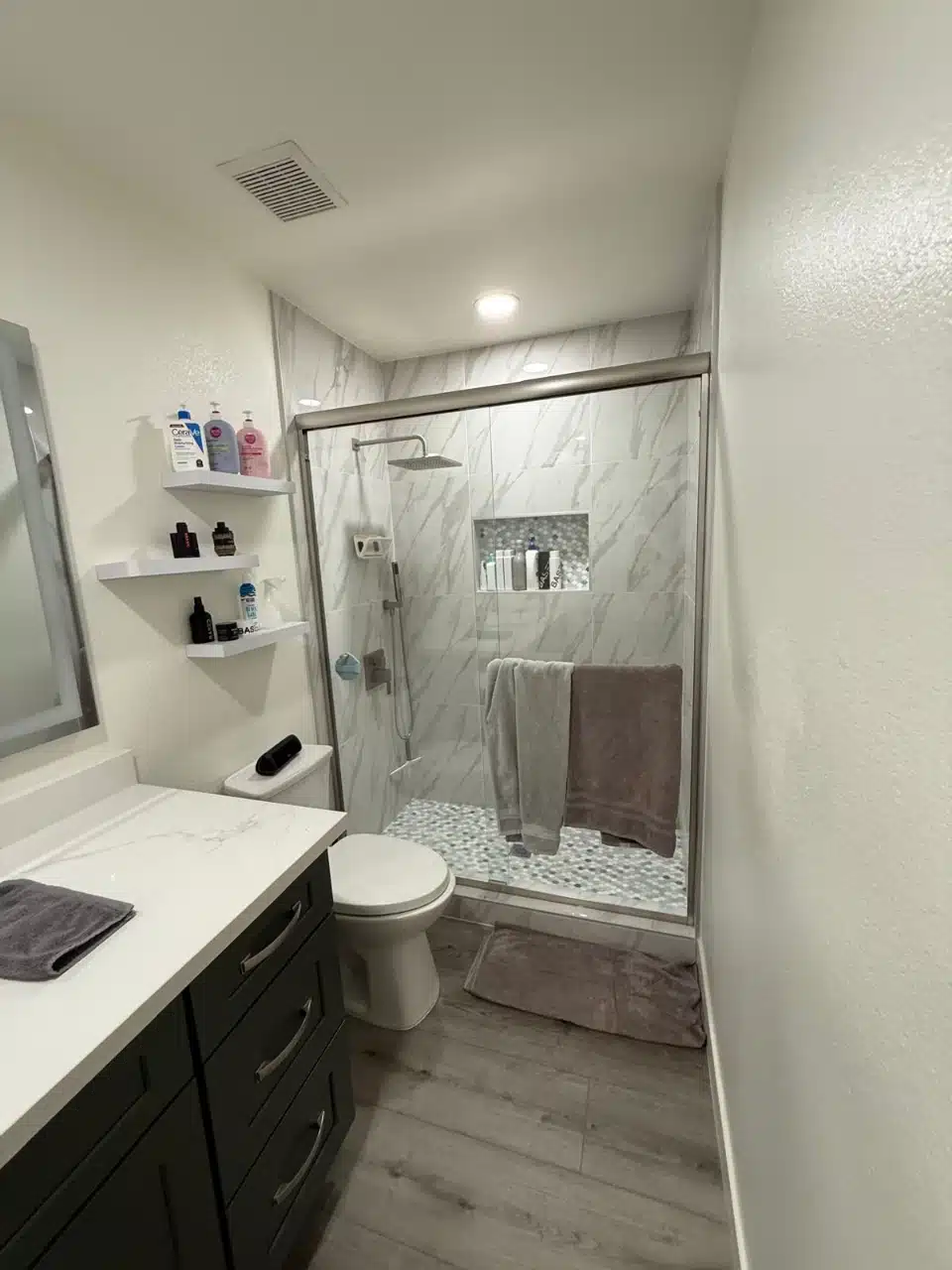 Second remodeled bathroom with glass shower, marble look tile, recessed niche, floating shelves and charcoal vanity