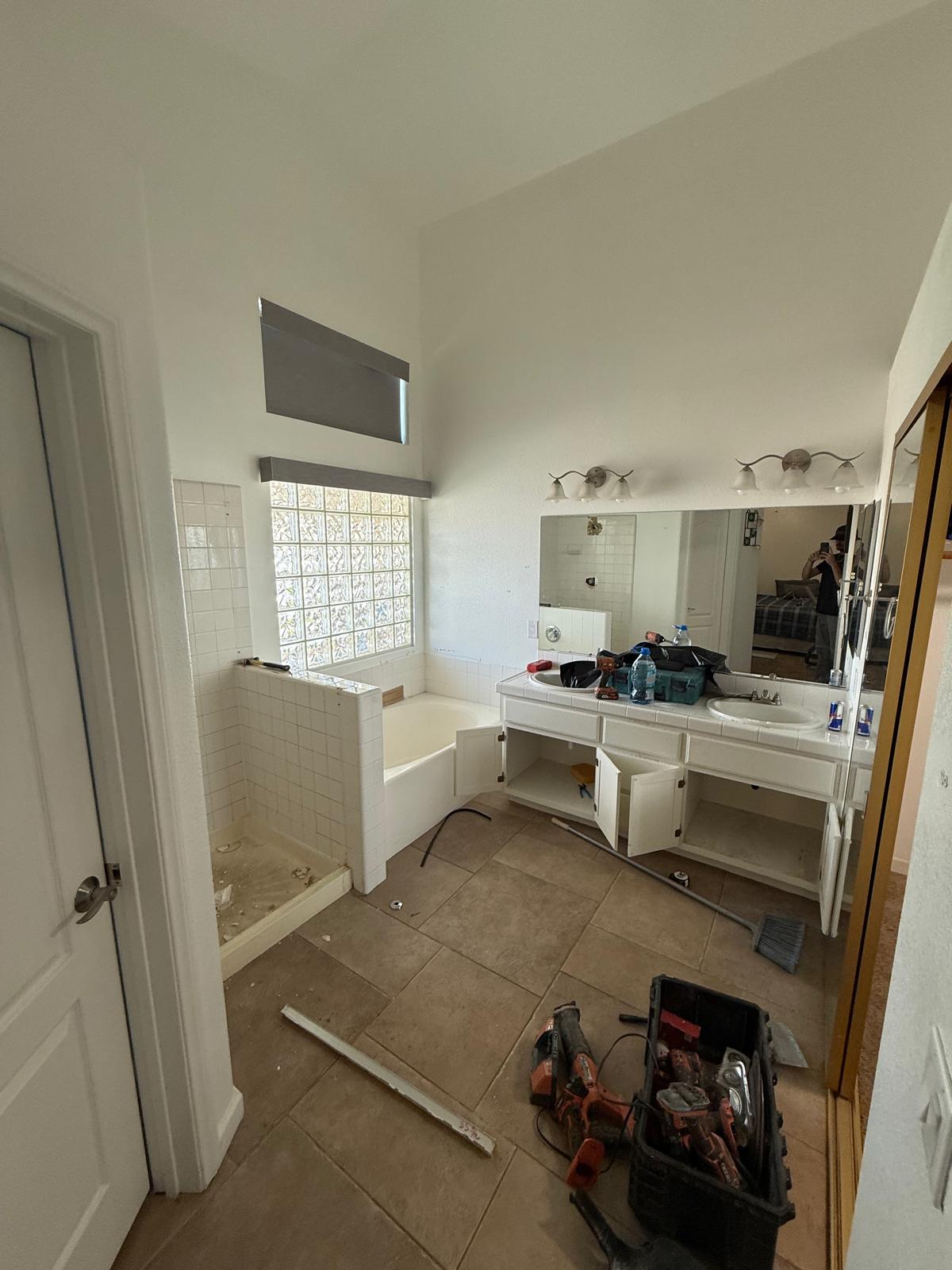 Bathroom during demolition with original floor removed, white tub surround, glass block window and white vanity with makeup counter