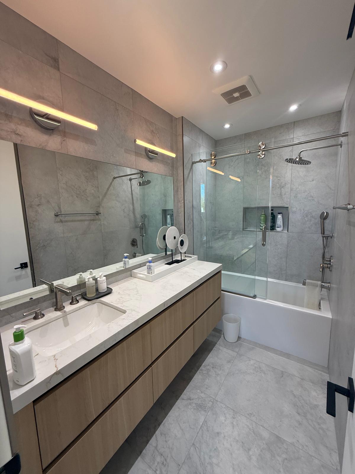 Modern primary bathroom with light oak floating double vanity, white countertop, wide mirror with LED lighting, gray wall tile and glass enclosed tub and shower