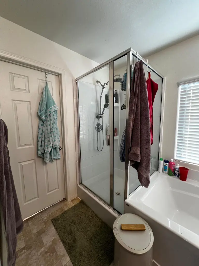Small bathroom with a glass shower enclosure, hanging towels, corner soaking tub, and bottles on the window ledge.