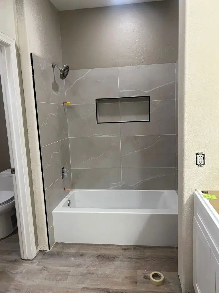 Tub and shower alcove with large gray wall tiles and a built-in niche during bathroom renovation.