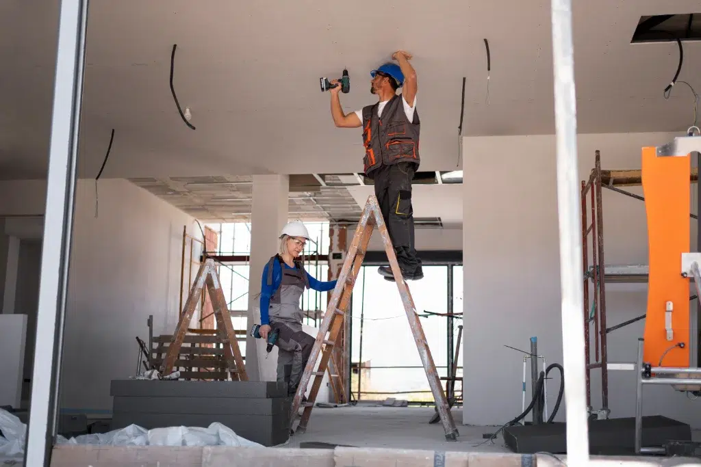 Construction workers on ladders installing ceiling panels with power tools inside a building under renovation.