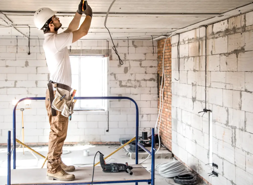 Construction worker standing on a scaffold installing electrical wiring in the ceiling of a partially finished interior space