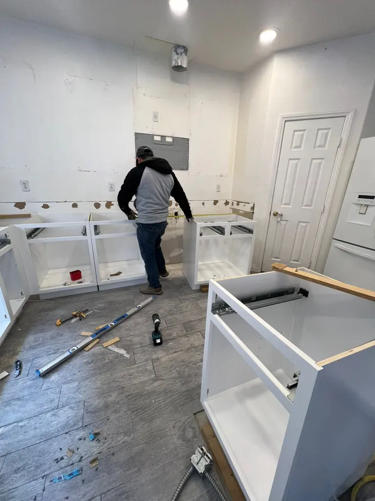 Kitchen renovation in progress with a worker installing white base cabinets, exposed wall areas, and tools scattered on the floor