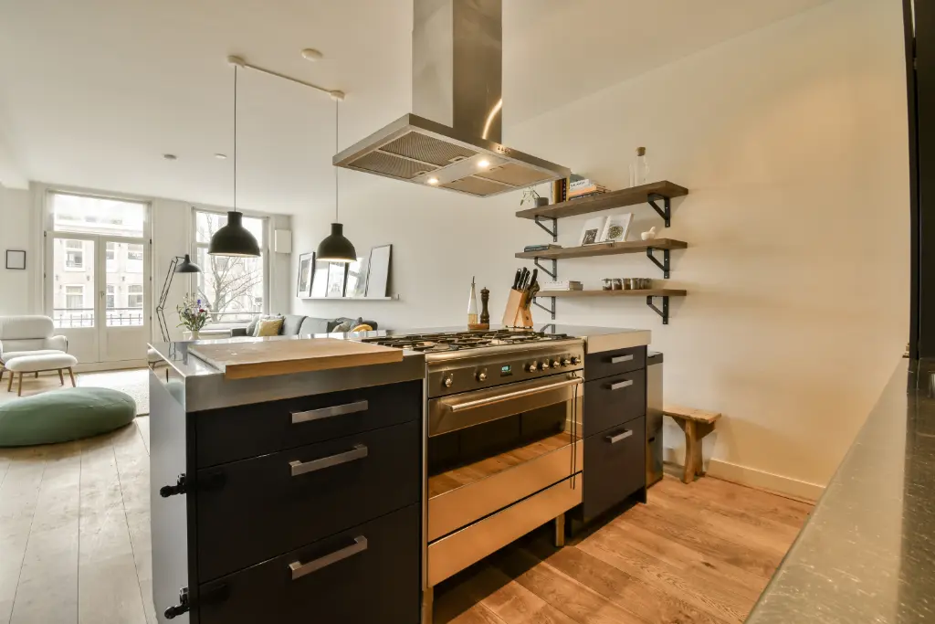 Modern open-plan kitchen with black cabinetry, stainless steel range and hood, wood floors, and floating shelves in a bright apartment